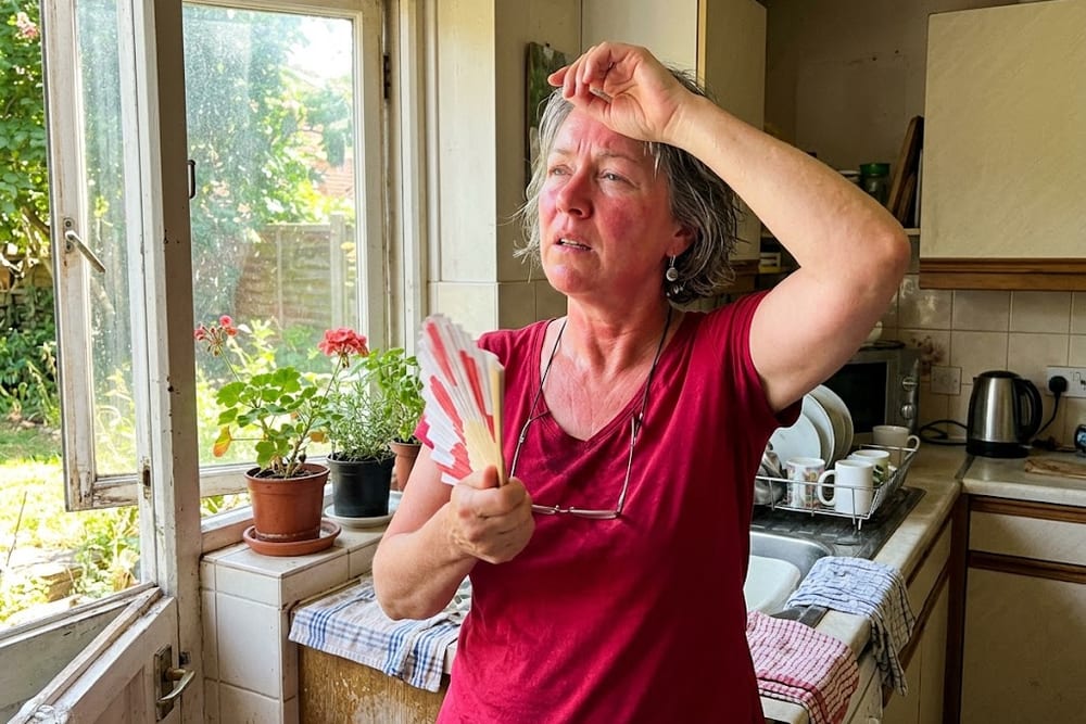 Woman age 50 in saturated cranberry red top fanning herself near a kitchen window during a hot flash moment, slightly flushed expression