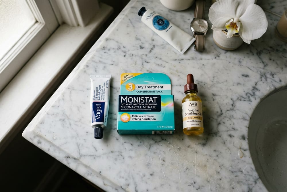 Overhead close-up of three different vaginal cream products lined up on a marble bathroom counter — one pharmaceutical tube, one turquoise OTC tube, one glass dropper bottle
