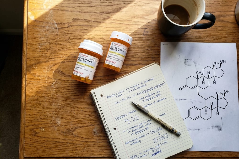 Overhead view of a desk with two amber prescription bottles, a printed molecular diagram showing two hexagonal hormone structures side by side, and an open notebook with handwritten notes