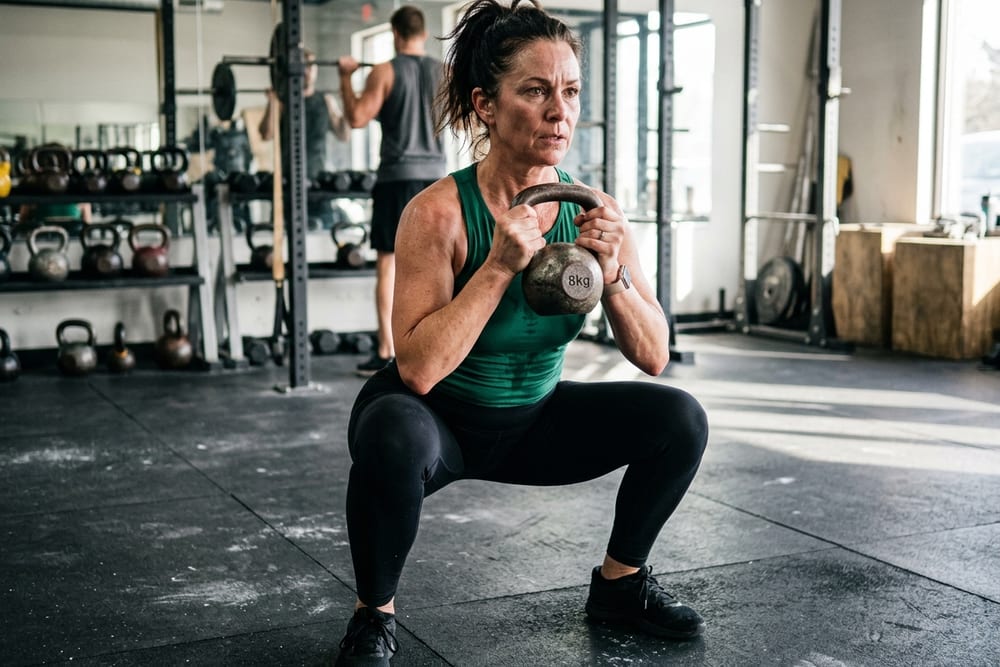 Woman age 49 in deep emerald green workout top doing a strength training squat with a light kettlebell on a rubber gym floor in morning light