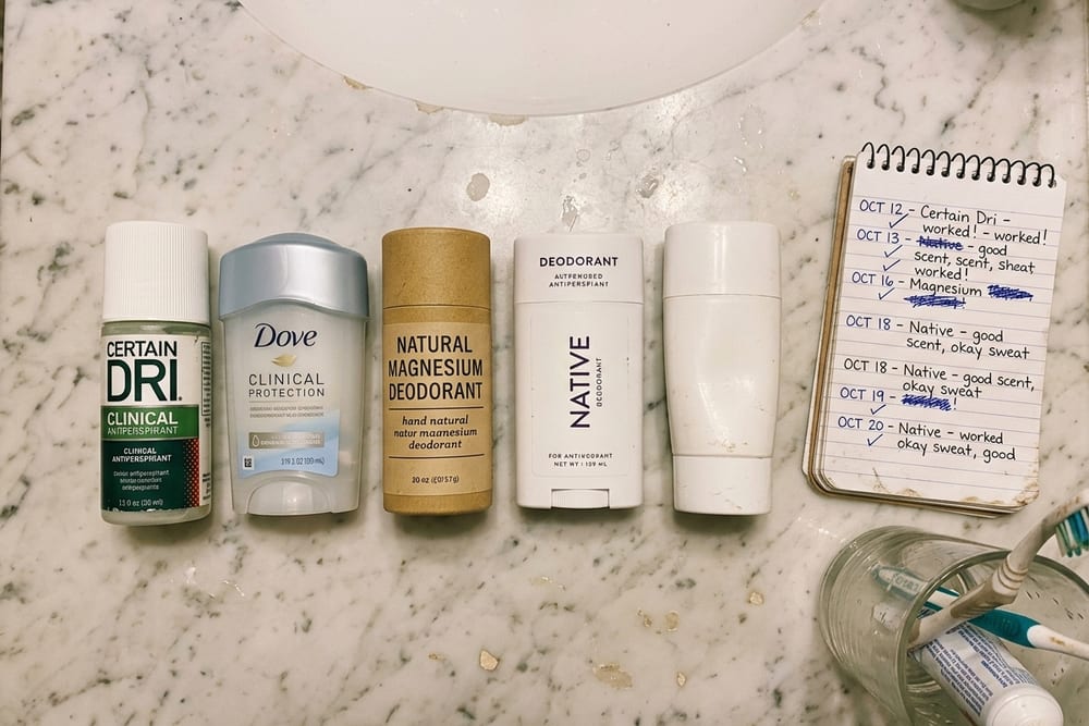 Extreme close-up overhead of a bathroom counter with five different deodorant tubes lined up alongside a small notebook with handwritten test notes in soft warm light
