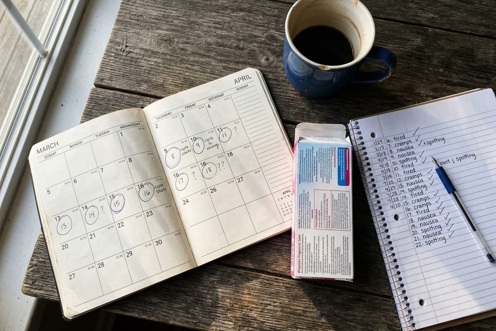 Overhead close-up of a kitchen table with a calendar showing several months of irregular cycle marks, a pregnancy test box, and a journal in real morning shadows