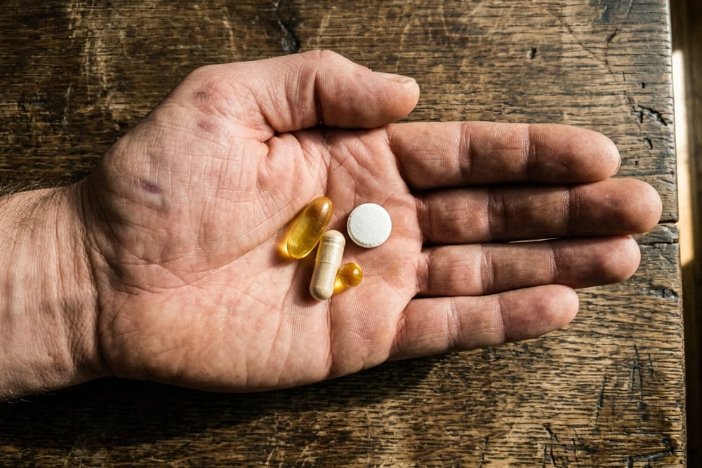 Open palm holding four different supplement capsules and tablets in directional warm window light on a weathered wooden table